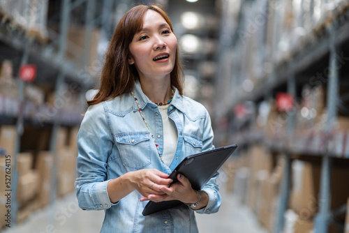Asian female employee counts the number of items on the shelf and holds a tablet to collect data to check the quality of the products in the warehouse.