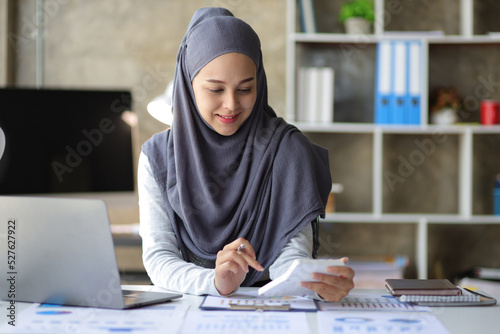 Attractive Muslim businesswoman working with laptop and analyzing financial statements on desk in office.