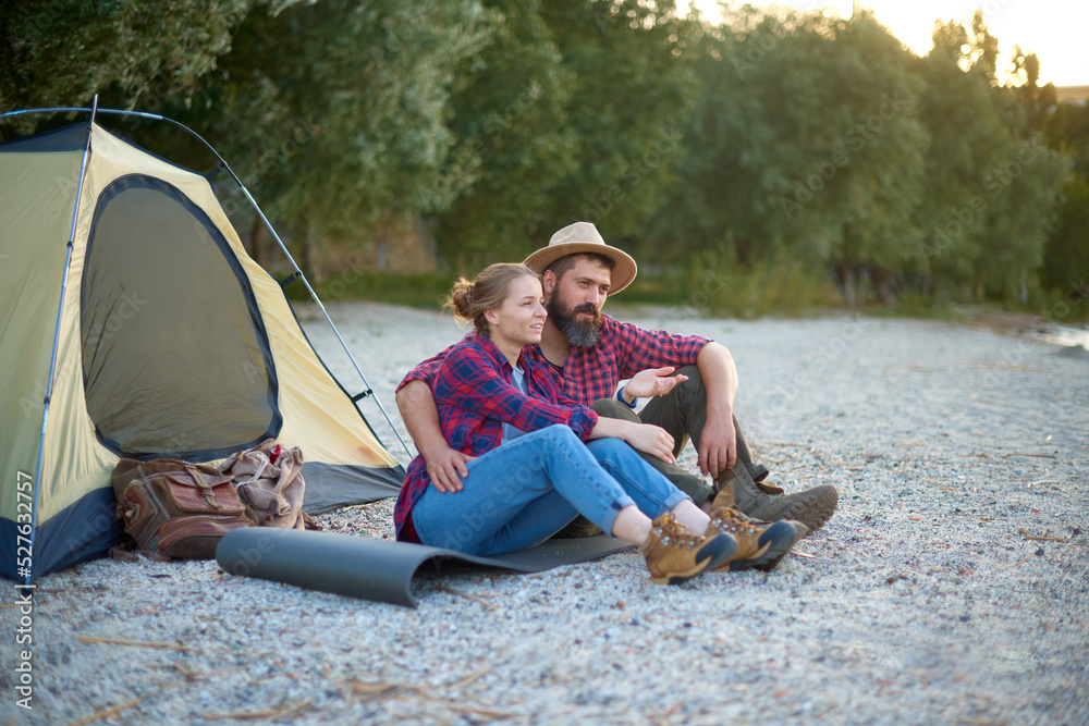 relaxing couple of woman and man sit near tent in camping, hugs, spend time together in weekend