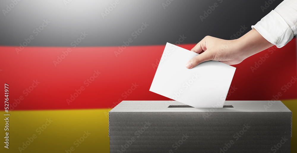 Close-up hand holding ballot paper into the voting box an Germany flag background. Freedom democracy concept. Freedom vote concept. Close-up hand holding ballot paper into the voting box an Germany flag background. Freedom democracy concept. Freedom vote concept.