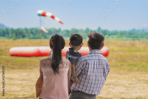 Back view of happiness Asian grandfather holding grandson with his daughter stands beside together in meadow with blurred image of cushion and wind sock in background. Happy family relationship.