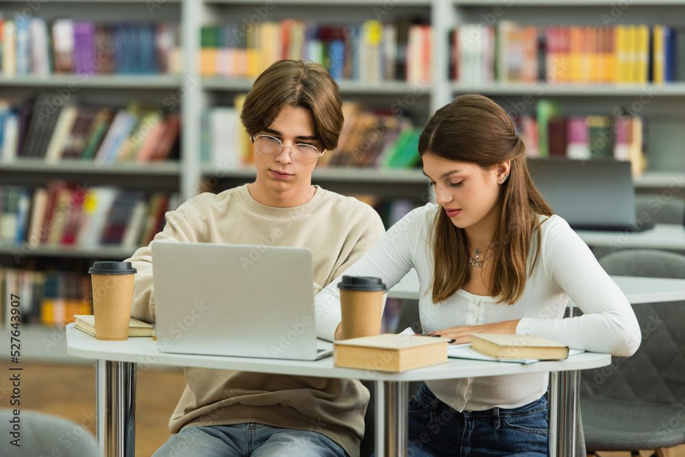 teenage students studying near laptop and paper cups in blurred library. teenage students studying near laptop and paper cups in blurred library.