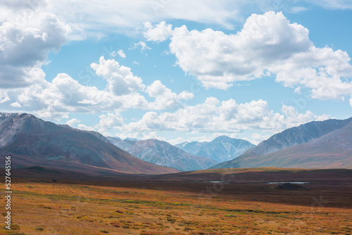 Motley autumn landscape of plateau with small lakes with view to sunlit mountain range under clouds in blue sky. Vivid autumn colors in mountains. Sunlight and shadows of clouds in changeable weather.