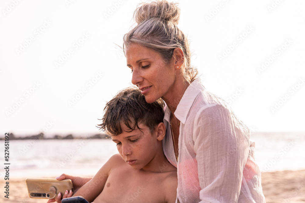 beautiful middle aged woman with her son using phone in relaxing sitting at the beach beautiful middle aged woman with her son using phone in relaxing sitting at the beach