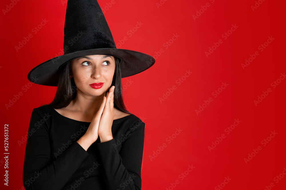Halloween. A young beautiful woman in a witch's hat and a black dress on a red colored background. Space for text. Halloween. A young beautiful woman in a witch's hat and a black dress on a red colored background. Space for text.