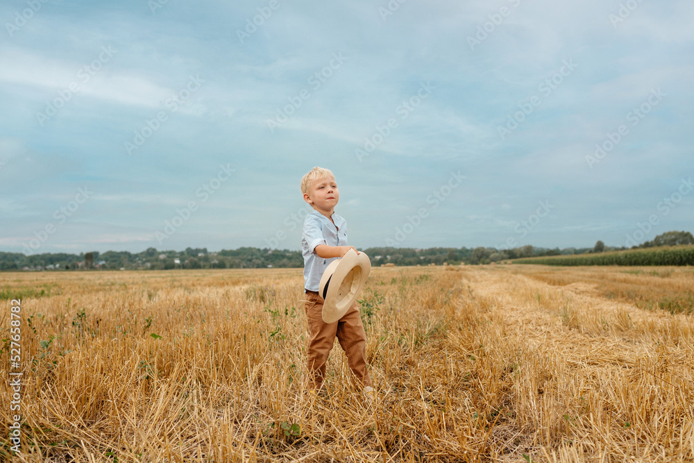 The blond boy is having fun playing with a straw hat in the summer in the field. The boy throws his hat into the sky. A happy child. Summer. Rural area