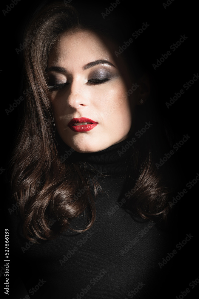 Beauty and fashion concept. Beautiful woman portrait standing in darkness with illuminated face. Model with evening makeup and red lips with closed eyes and wavy hair. Low key photography style Beauty and fashion concept. Beautiful woman portrait standing in darkness with illuminated face. Model with evening makeup and red lips with closed eyes and wavy hair. Low key photography style