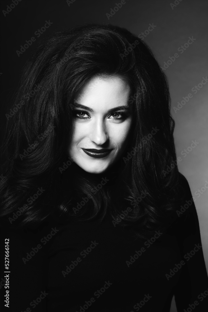 Beautiful woman with long dark fluffy and wavy hair studio portrait. Model with evening make-up wearing dark clothes in dark studio background looking to camera. Black and white image