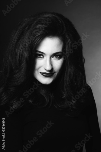 Beautiful woman with long dark fluffy and wavy hair studio portrait. Model with evening make-up wearing dark clothes in dark studio background looking to camera. Black and white image