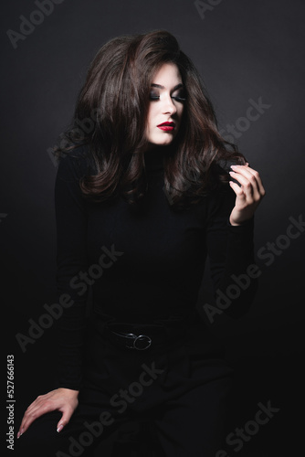 Beautiful brunette woman with long dark fluffy and wavy hair studio portrait. Model with evening make-up and red lipstick wearing dark clothes in dark studio background looking to her nails