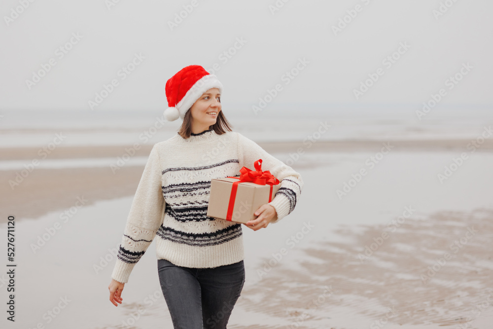 Obraz premium woman in a Santa Claus hat with a gift box in her hands smiles. a happy European woman carries a gift for New Year or Christmas. give gifts and surprises, poster postcard. holiday holidays Christmas