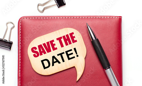 On a light background there are black paper clips, a pen, a burgundy notepad a wooden board with the text SAVE THE DATE