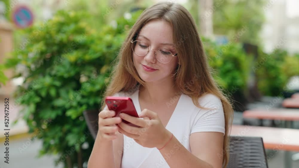 Young blonde woman using smartphone sitting on table at coffee terrace