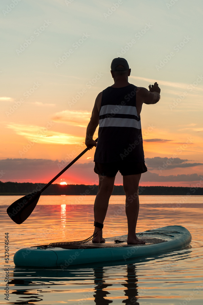 A man in shorts stands on a SUP board with an oar at sunset in a lake against the backdrop of a sunset sky.