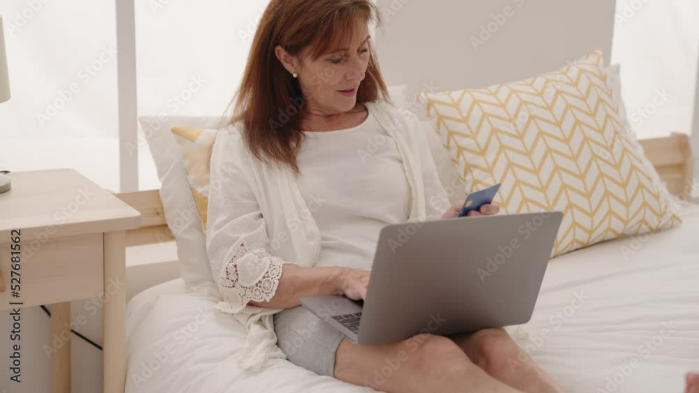 Middle age woman using laptop and credit card sitting on bed at bedroom Middle age woman using laptop and credit card sitting on bed at bedroom