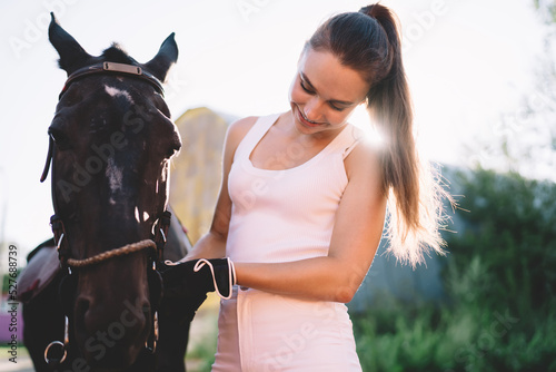 Positive young horsewoman tying bridle and smiling in countryside