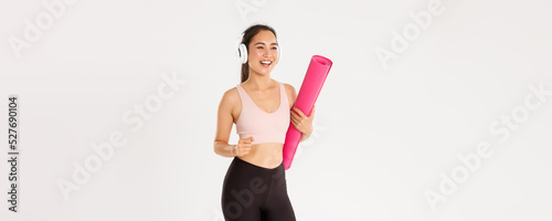 Sport, wellbeing and active lifestyle concept. Cheerful smiling asian fitness girl in headphones and sportswear going to gym with rubbermat, hurry up for yoga classes excited, white background photo