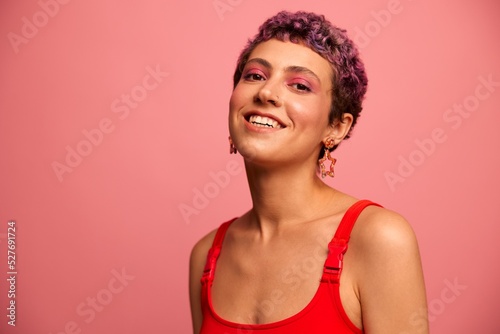 Fashion portrait of a woman with a short haircut of purple color and a smile with teeth in a red top on a pink background happiness