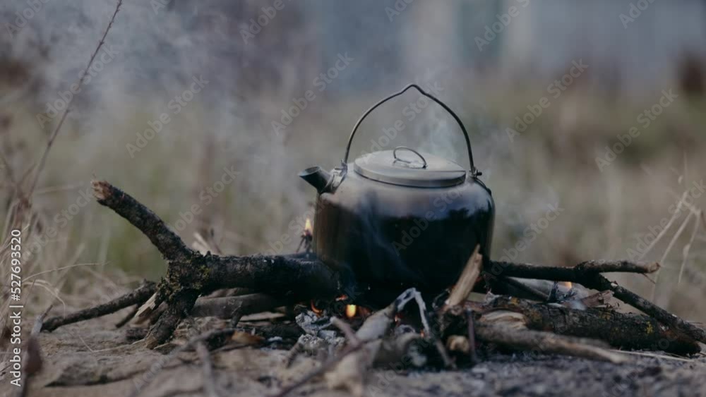 A campfire on the grass during a hike is lit by hikers and they boil water in an iron kettle to make tea to get warm A campfire on the grass during a hike is lit by hikers and they boil water in an iron kettle to make tea to get warm