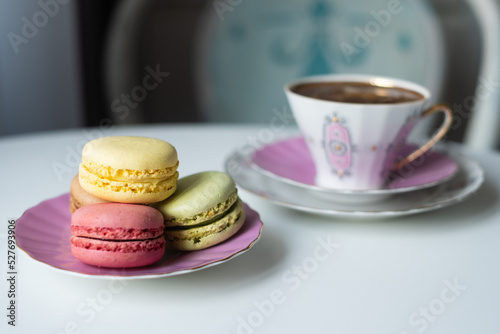 Mix of appetizing macarons and delicious coffee in porcelain cup on white table decorated with blurred flowers. Dry flowers. Greeting card.