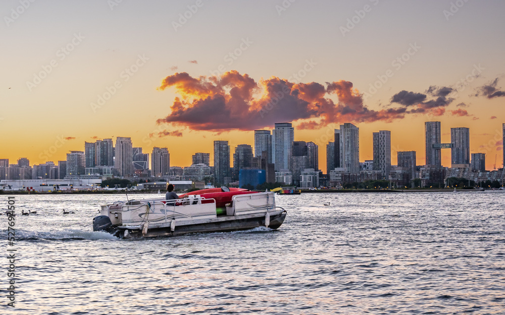 Fototapeta premium A pontoon boat loaded with colorful kayaks heads across Toronto's Inner Harbour with the city skyline, and airport and the sunset in the background.