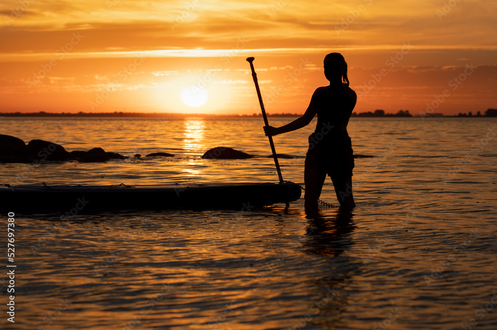 silhouette of a teenage girl standing in the water next to a Stand Up Paddle Board SUP, holding the paddle in her hand.  Golden sunset sky reflecting in the water. Greifswalder Bodden, Baltic Sea