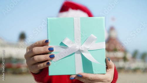 Smiling young woman in red Santa Claus hat smiling giving elegant stylish teal blue Christmas gift box with white satin ribbon outdoors on tropical beach. Happy female with present. Winter holidays US photo