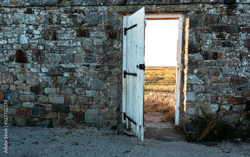 Canvas Print Old, dark, stone wall with white, vintage, wooden exit door overlooking bright sunny meadow - landscape