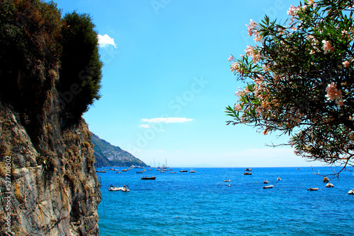 Enchanting view in Positano with a big slab of rock on the left, a tree on the right, the colorful Tyrrhenian Sea filled with many boats in the middle and a mountain in the background