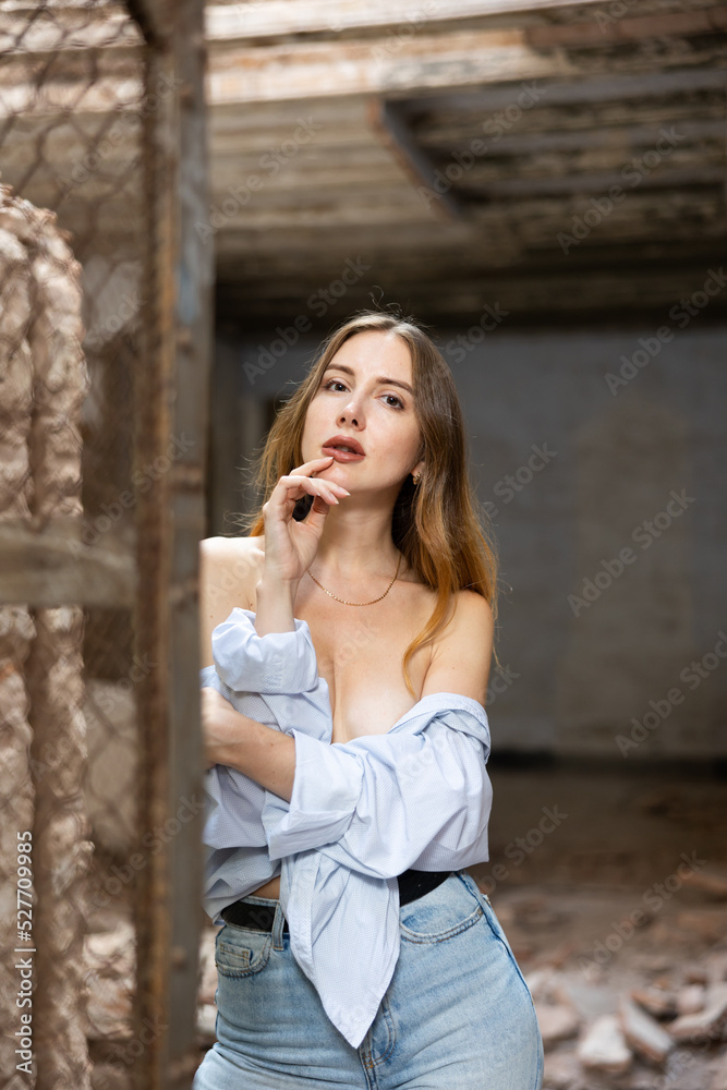 Young woman in blue jeans and open shirt slightly revealing her breast posing sensually during fashion photo shoot on grunge background near steel mesh fencing 