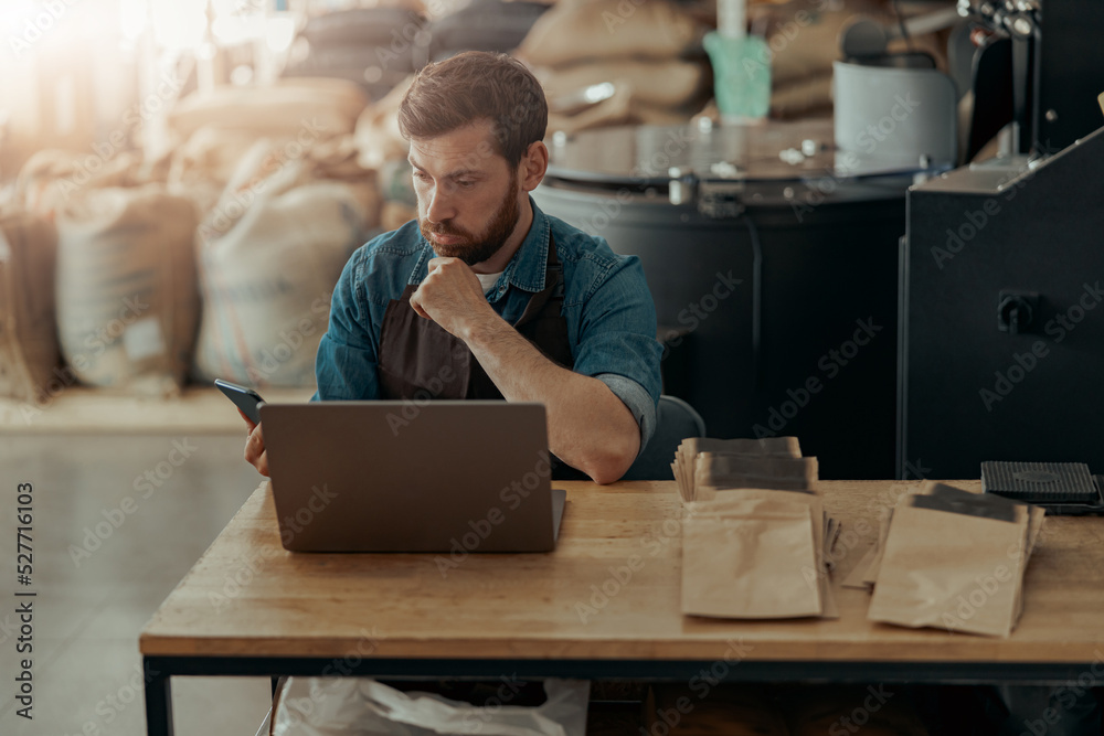 Business owner of small coffee roasting factory working on his workplace using laptop and phone Business owner of small coffee roasting factory working on his workplace using laptop and phone