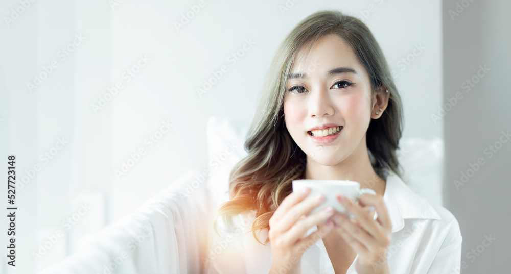 Cropped view of Cute Young Asian woman drinking coffee from cup at morning, Woman in white pajama sitting on bed and drinking coffee at the sunny morning bedroom. Cropped view of Cute Young Asian woman drinking coffee from cup at morning, Woman in white pajama sitting on bed and drinking coffee at the sunny morning bedroom.
