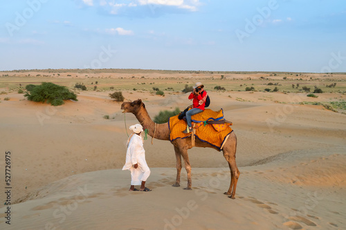 Female tourist taking picture while riding camel, Camelus dromedarius, at sand dunes of Thar desert, Rajasthan, India. Camel riding is a favourite activity amongst all tourists visiting here. © mitrarudra
