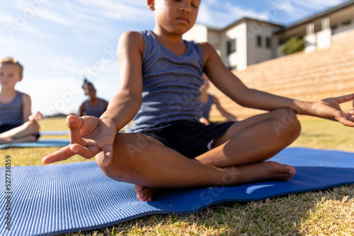 Biracial elementary schoolboy gesturing while meditating on exercise mat during sunny day