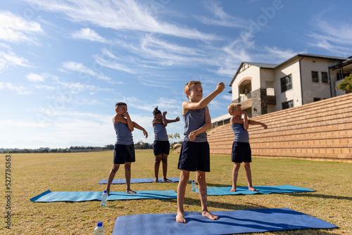 Full length of multiracial elementary schoolboys doing stretching exercise on yoga mat against sky
