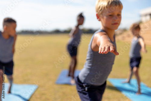 Multiracial elementary schoolboys doing warrior 2 pose while exercising on yoga mat during sunny day