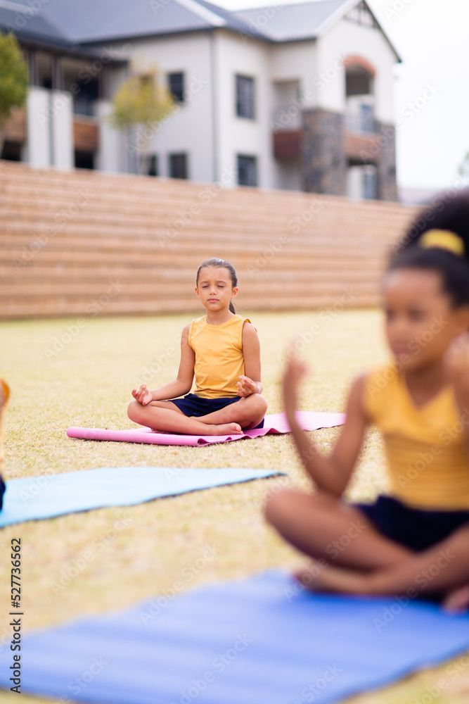 Fototapeta premium Multiracial elementary schoolgirls meditating while sitting on yoga mat against school