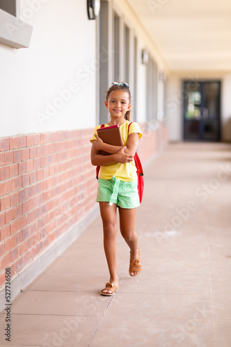 Full length portrait of caucasian elementary schoolgirl with books and backpack walking in corridor
