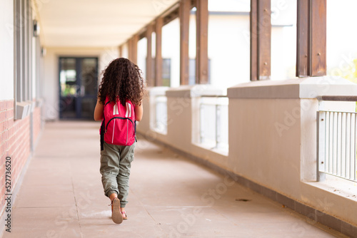 Rear view full length of biracial elementary schoolgirl with backpack walking in corridor at school