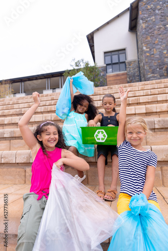 Happy multiracial elementary schoolgirls with garbage bags sitting on school steps