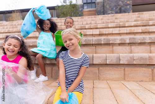 Cheerful multiracial elementary schoolgirls with garbage bags sitting on school steps