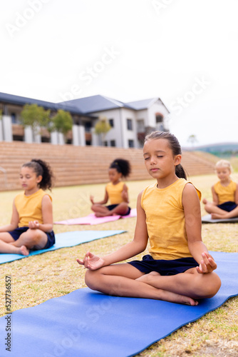 Multiracial elementary schoolgirls meditating while sitting on exercise mat at school ground