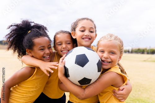 Happy multiracial elementary schoolgirls with soccer ball embracing while standing on ground