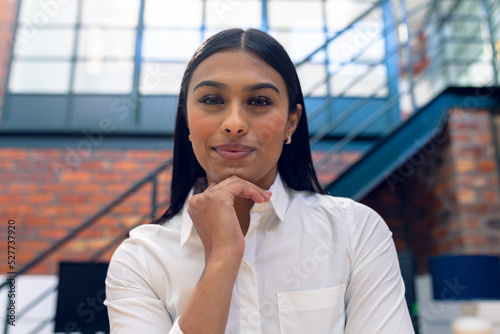 Portrait of smiling biracial young businesswoman with hand on chin sitting in office