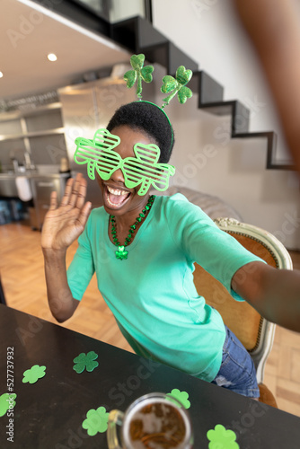 Happy african american woman wearing shamrock novelty glasses while taking selfie at home