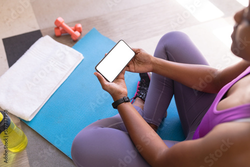 African american mid adult woman using smart phone while sitting on exercise mat at home, copy space