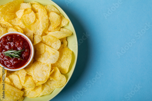 Red sauce in bowl amidst potato chips served in plate on blue background with copy space