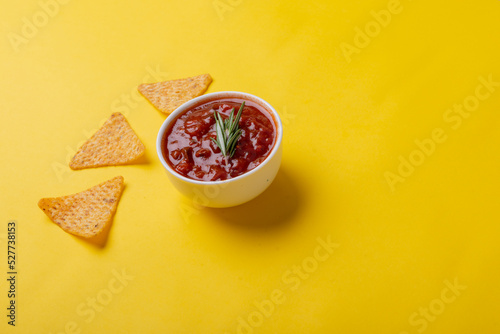 Red sauce with rosemary served in bowl with nacho chips on yellow background with copy space