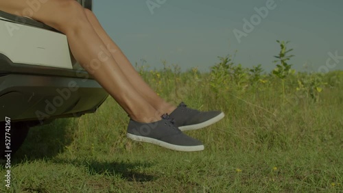 Close-up of slim beautiful female legs in canvas shoes dangling from car trunk over scenic summer nature, while woman traveler enjoying road trip and vacations in countryside.