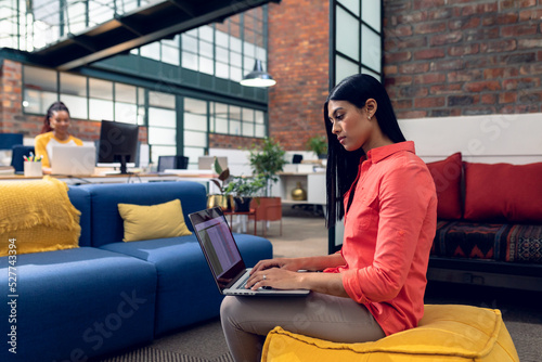 Biracial young businesswoman using laptop while sitting on ottoman in modern office photo